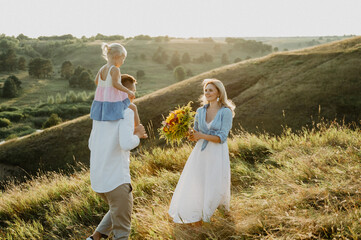 A young family with a little daughter are hugging and walking in the field at sunset.