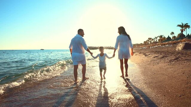 Back view happy family holding hands and together walks barefoot along seashore in water waves with foam near beach. Summer landscape nature outdoors. Sport lifestyle on travel leisure activity.