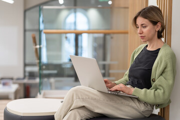 Focused pensive middle-aged woman sitting on ottoman with laptop, scandinavian lady freelance...