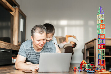 Dad works at home on a laptop and plays with his son at the same time. The child hugs dad and...