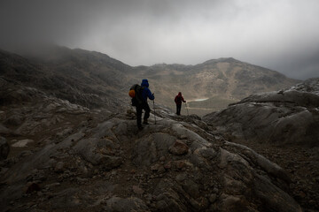 two men hiking toward a mountain hut in overcast bad weather amidst a barren mountain landscape with dramatic lighting conditions