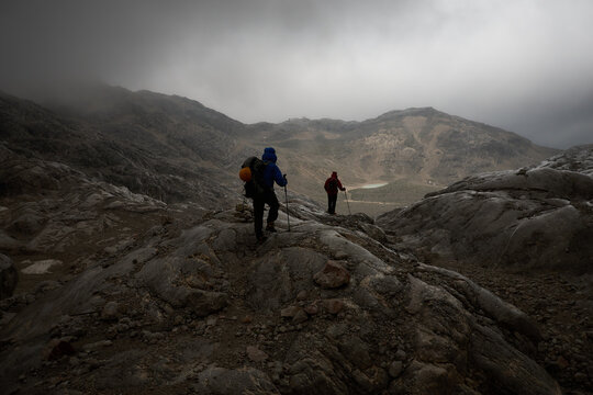 Two Men Hiking Toward A Mountain Hut In Overcast Bad Weather Amidst A Barren Mountain Landscape With Dramatic Lighting Conditions