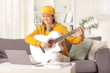 Young female with an acoustic guitar and headphones sitting in front of a laptop computer