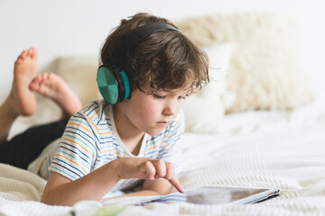 Cute boy lying on the bed at home and playing with pc tablet or reading online and listening to...