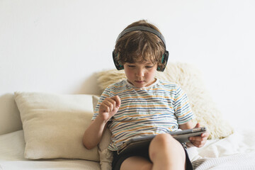 Cute boy lying on the bed at home and playing with pc tablet or reading online and listening to music with wireless headphones