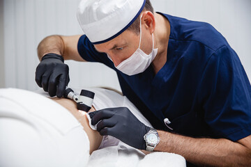 A cosmetologist male doctor in black gloves and blue uniform examines the patient's mole using a dermatoscope. 