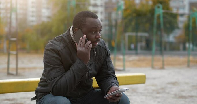 African American Man Sits At Sport Ground, Speaks On Smartphone And Holds In Hand Second Smartphone. Cinema 4K 60fps Video
