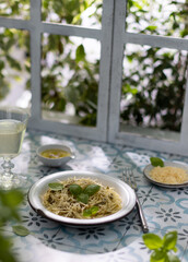 Spaghetti with pesto sauce,fresh basil,cheese,glass of white wine on blue tiles near the wooden window.Italian dish on summer table.