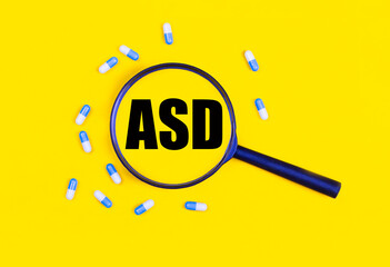 Pharmaceutical tablets and capsules near a magnifying glass with the text ASD Autism Spectrum Disorder on a yellow background. View from above.