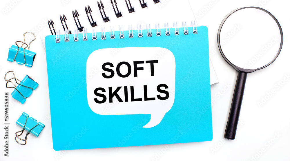 Poster office table with blue notepad, paper clips, magnifying glass and white card with text soft skills. 