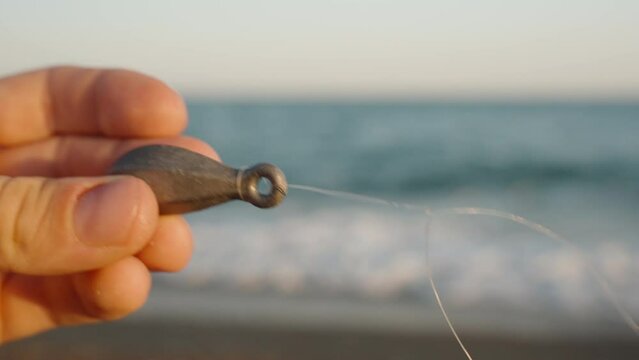 A male angler shows a tackle for fishing at sea, close-up. Sinker and hook. Sea and waves in the background.