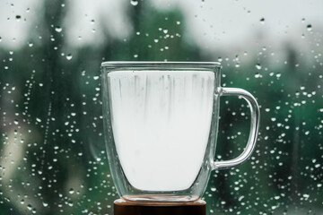 Glass cup filled with water on nature background and window. Transparency cup with water, near window with drops after rain and green blurry background.