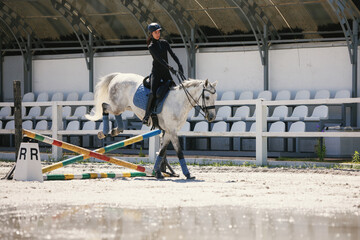 Portrait of young woman in sports uniform and helmet training at riding arena in summer day,...