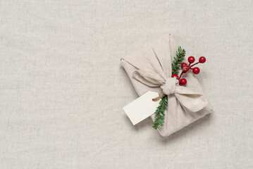 Fototapeta na wymiar Christmas gift wrapped in fabric with decorative holly, fir branch and blank label. Beige linen textile background. Traditional Japanese Furoshiki gift. Zero waste concept. Top view, flat lay. 