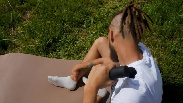 Concept healthy lifestyle. Caucasian man with dreadlocks sits on mat in park and kneads muscles of shoulders and neck after training using percussion massage gun. 4K slow motion horizontal footage.