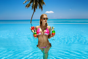 Beautiful female model in pink bikini is standing in the pool and holding colorful fruity drink....