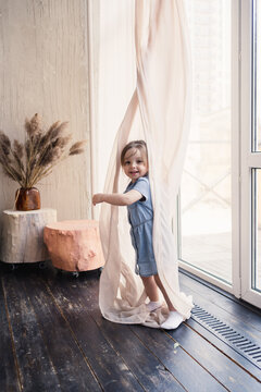 A portrait of a happy child, cute little baby or toddler girl hiding behind the curtain near the glass window. High quality photo