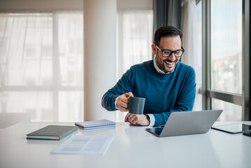 Cheerful professional enjoying coffee while working on laptop at office desk.