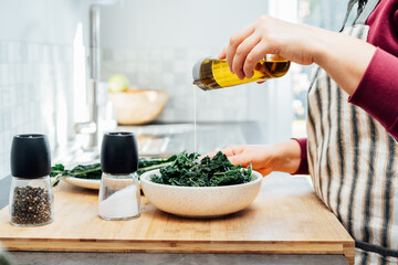 Close up woman adding olive oil while cooking kale chips or healthy salad for dinner on the kitchen...