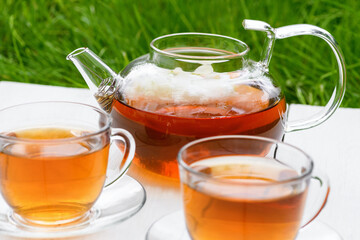 Tea in glass teapot and two cups on a wooden white background background against the background of green grass. Teapot with green or black tea. Tea time