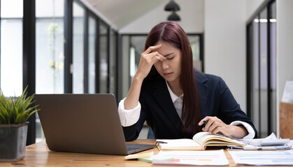 Obraz premium Young Asian woman feeling migraine head strain. Tired, Overworked businesswoman financier while working on laptop computer in office.