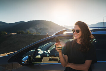 Young beautiful woman traveling by car in the mountains using smartphone at sunset, summer vacation and adventure