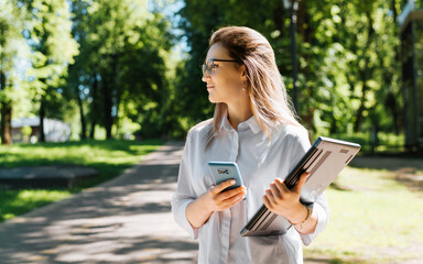 Half-length portrait of young female student or frilnaser with laptop and smartphone standing in park and looking away. Pretty cute young woman wearing glasses and white shirt outdoors.