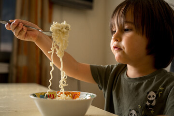 a boy with long dark hair is sitting at home in the kitchen and eating long Chinese noodles with a...