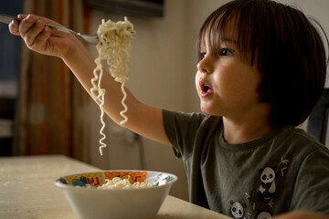 a boy with long dark hair is sitting at home in the kitchen and eating long Chinese noodles with a fork. A child eats instant noodles