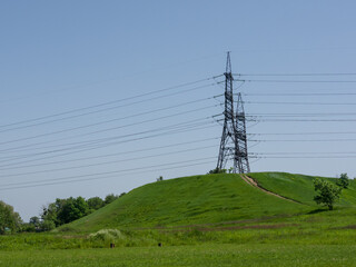 High-voltage towers with wires on a hill in the countryside, among grass and bushes, on a sunny summer day.
