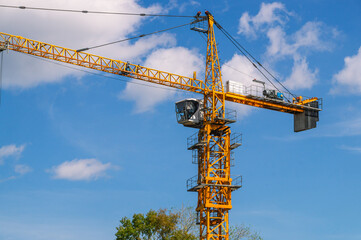 A tower construction crane on the background of a blue sky with clouds. Boom rotary crane with boom. Construction of apartment buildings in the city. Tower crane cabin.