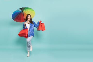Young Asian woman wearing raincoat and holding rainbow umbrella and shopping bag isolated on green background, Shopaholic in the rainy season concept © comzeal
