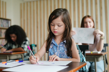 Preschool kids drawing with color pencil on white paper on table in classroom with friends, kindergarten education concept