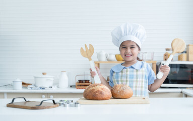 Portrait of Asian little cute kid girl wear chef hat holding wooden fork and spoon, smile at camera with fresh loaf of bread in kitchen at home or cooking class at school. Child education concept