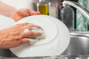 Young woman's hands washing a white dish with water and dish soap. Close-up of female hands rinsing a dirty and used dish with the faucet turned off. Concept of cleanliness