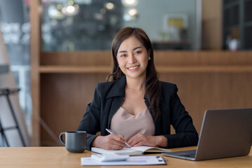 Smiling beautiful Asian businesswoman sitting at work looking at camera using laptop and computer...