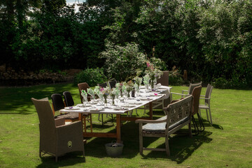 Large wedding table with crockery and flowers served in the garden on green grass. Summer holiday in the garden by the pond. Festive table with treats, glasses, decorated by a florist with herbs 