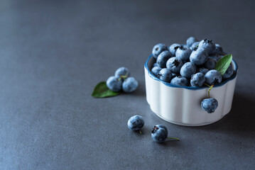 Ripe blueberry in a clay white bowl and a few berries nearby on the dark grey background. Copy space. Fitness conept. Healthy nutrition