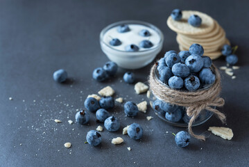 Fresh blueberry and yogurt in the glass jars, cookies and scattered blueberries on the dark grey background. Healthy nutrition