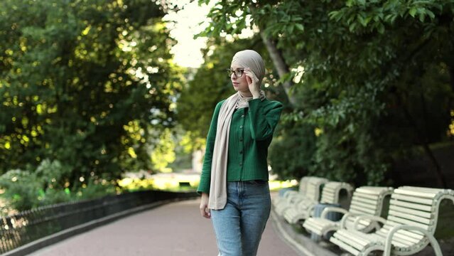 Smiling young muslim woman touching her eyeglass during break outdoor. Cheerful arabic woman in hijab walking in green summer city park.