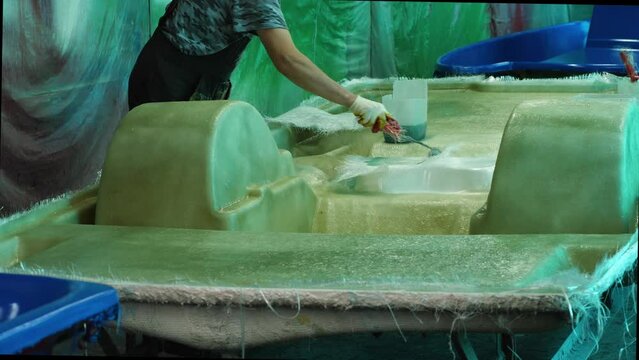 A man uses a paint roller to apply adhesive to fiberglass, gluing the hull of a pedal catamaran. Building boats in a fiberglass shipyard