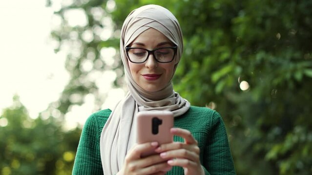 Portrait of beautiful smiling stylish muslim businesswoman with smart phone in hijab walking in summer park. Outdoor, park, business, gadget concept. Walking down the sunny city park.