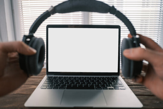 Desktop with modern silver laptop with white screen for mockup. Point of view. A man puts on headphones.