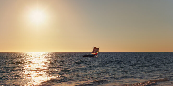 Sun setting down to calm sea, small sail piroga boat with silhouette of unknown man in distance