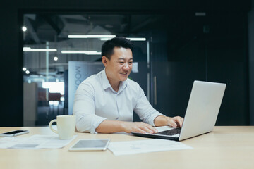 Young handsome male Asian freelancer conducts a video meeting on a video call from a laptop. Sitting at the table in a modern office, smiling, talking.