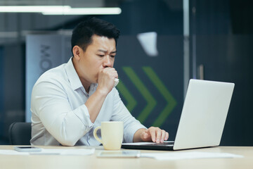 Fototapeta na wymiar Sick at work. A young Asian businessman keeps coughing, covers his mouth at the desk in the office, feels bad, has an allergy, feels chest pain, needs help.