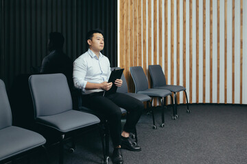 A young handsome Asian man is waiting in the reception hall for an interview for a job in an office, bank, business company. He holds a folder in his hands, worries, shakes it off