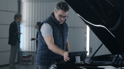 Fototapeta na wymiar In a car service a male mechanic inspecting a car engine and making a diagnosis using a gadget while a female mechanic consulting a man