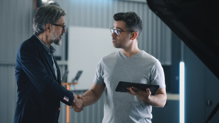 Manager in a suit with a tablet shaking hands with a male client after car diagnostics in a car service repair shop