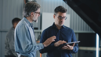 A manager in casual clothes and a male client standing near the car in a car service during diagnostics and watching data on a tablet